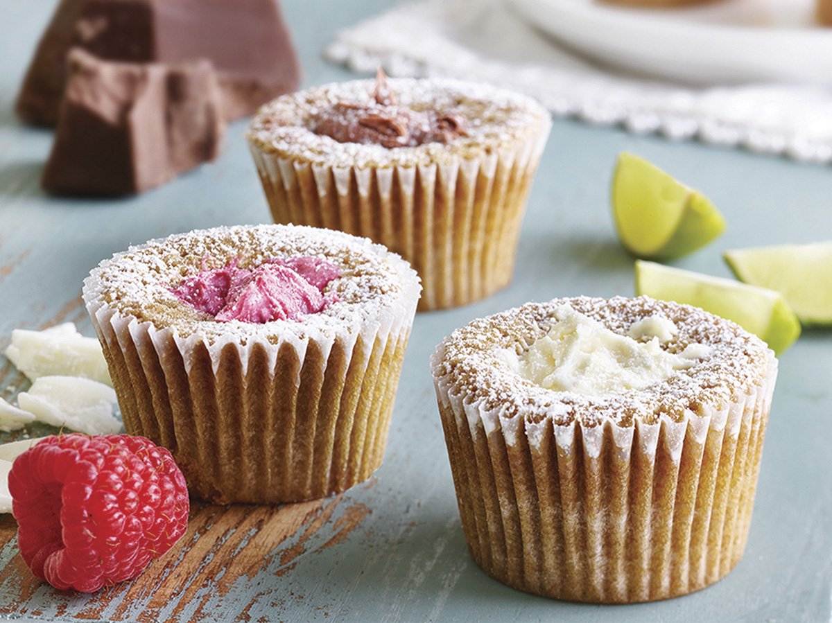 Three decorated cupcakes with different frosting flavors sit on a wooden surface, accompanied by fresh raspberries and lime wedges.